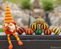 A crocheted gnome doll with a tall orange, yellow, and white striped hat sits on a dark wooden surface next to a small stack of crocheted pumpkins in orange, black, green, and yellow. The background is a softly blurred outdoor garden setting.