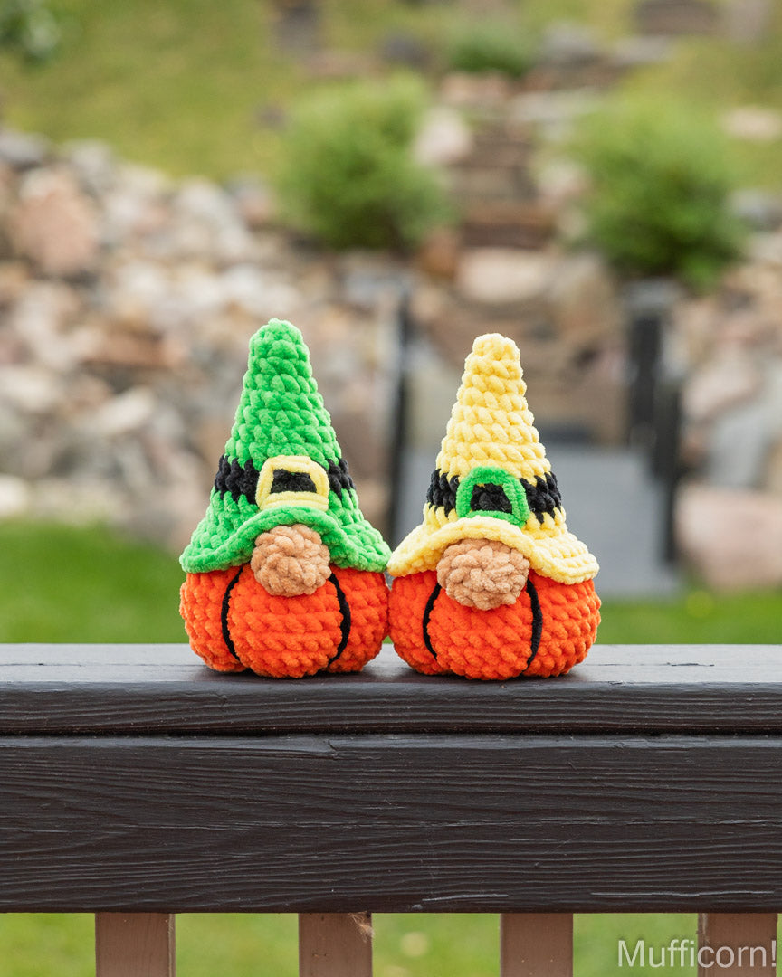 Two crocheted gnome pumpkins sitting side by side on a dark wooden railing, with a blurred outdoor garden background. One gnome has a green hat and the other has a yellow hat.