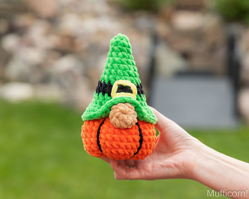 A hand holds a crocheted gnome pumpkin against a blurred outdoor background of green grass and a stone wall. The gnome has a green pointed hat with a black band and gold buckle detail, an orange pumpkin body with black stitched veins, and a brown nose.