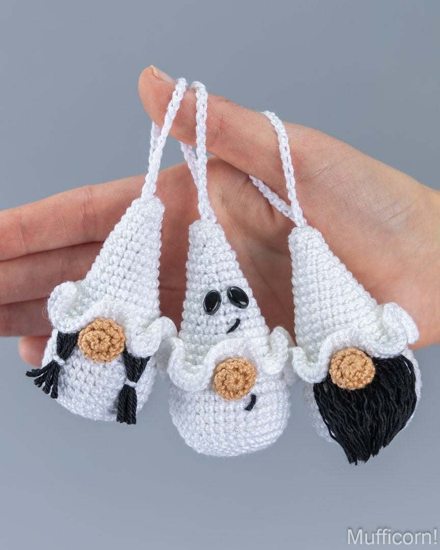 Three small crocheted white muffin-shaped ornaments with hanging loops are held in a person's hand against a plain gray background. The ornaments have black yarn hair, black yarn eyes, and tan yarn cheeks.