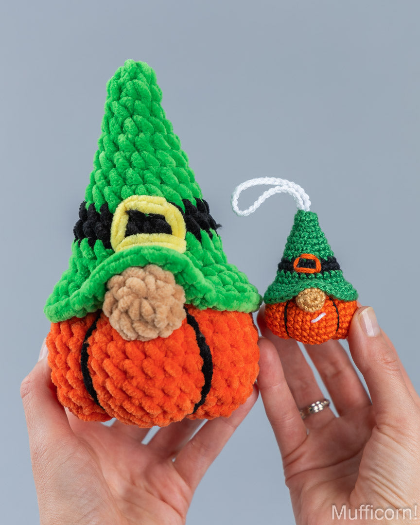 Two crocheted pumpkin gnomes held in hands against a plain gray background. The larger gnome is orange with a green hat, black and yellow details, and a beige beard.