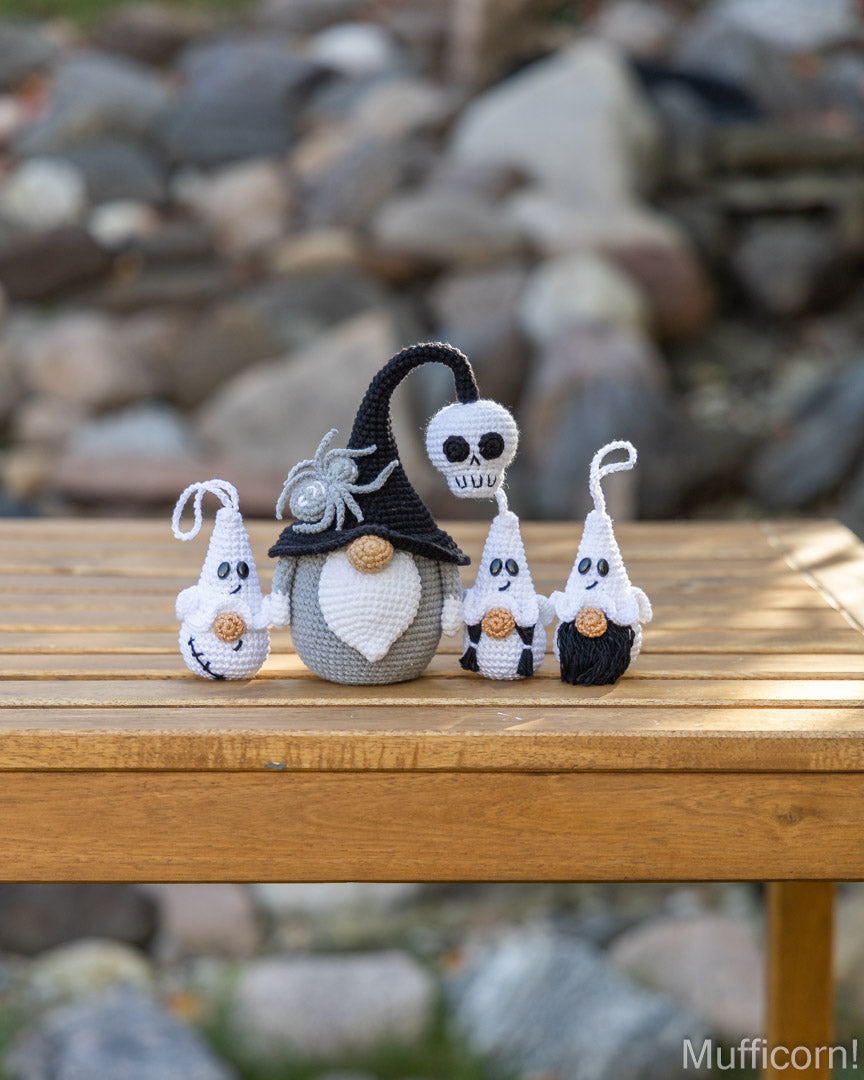 Three crocheted Halloween characters, including a witch with a large black hat, a skull, and two small ghosts, displayed on a wooden table with a blurred outdoor stone background.