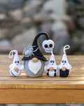 Four crocheted Halloween ornaments displayed on a wooden surface with a blurred outdoor stone background. The set includes a gray gnome with a black pointed hat and white beard, a white skull figure, two small white-and-black ghost figures, and two tiny white-and-black candy corn figures.