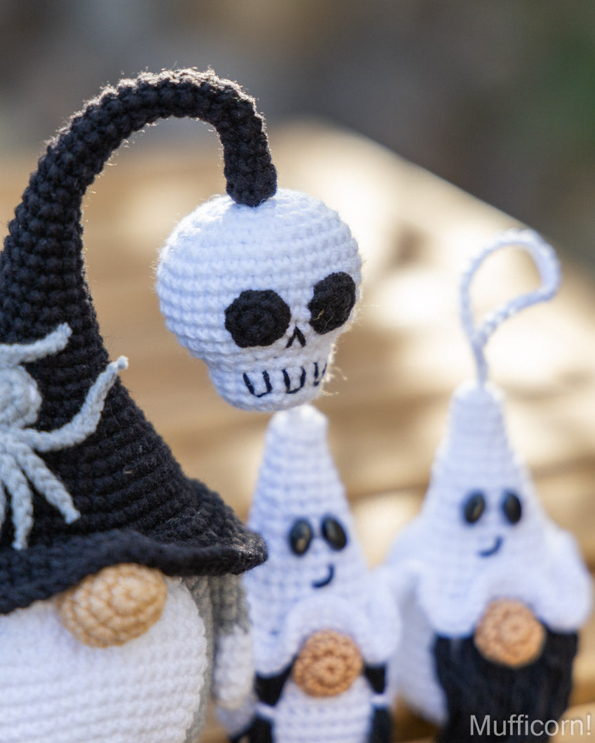 Close-up of crocheted Halloween ornaments, including a black witch hat with a white spider, a white skull, and ghost figures with candy accents, photographed outdoors with a blurred natural background.