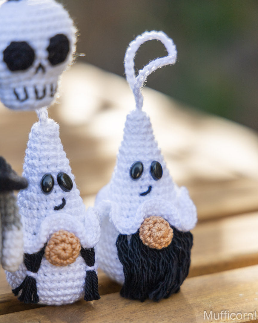 Three handmade crocheted ghost ornaments with hanging loops, displayed on a wooden surface outdoors. The ghosts are primarily white with black yarn details for eyes, mouths, and hair, and one holds a round tan cookie.