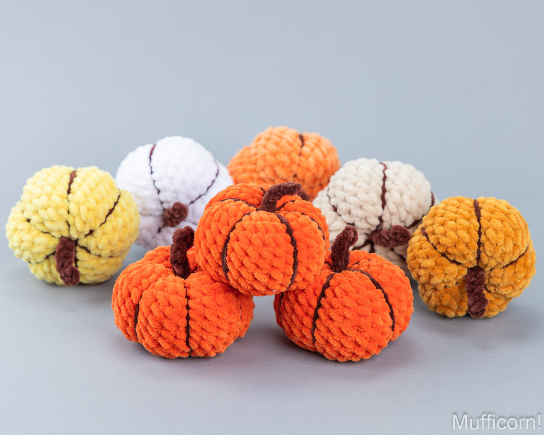 A set of crocheted pumpkin-shaped ornaments in orange, yellow, white, and cream, arranged on a smooth light gray surface with a soft gray background.