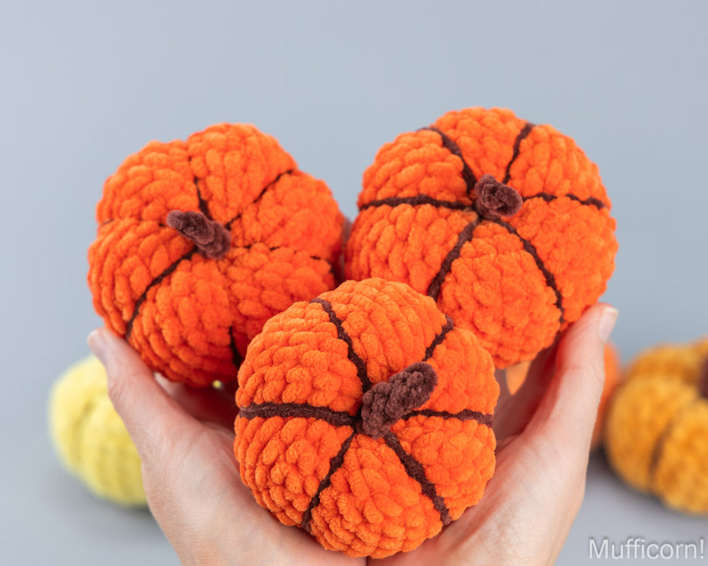 Three small orange yarn pumpkins with dark brown stitched seams and stems are held in a person's hands against a soft gray background.