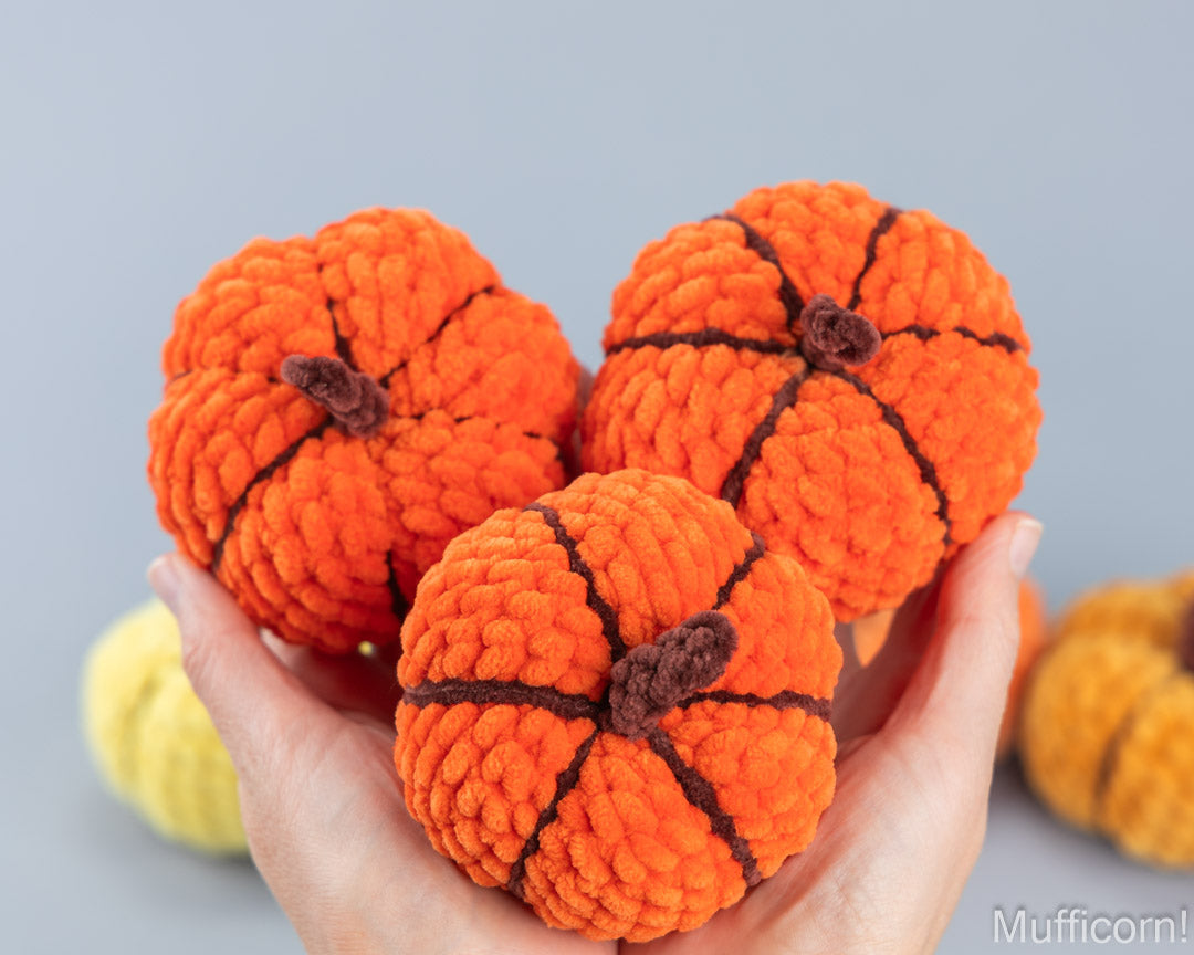 Three small orange yarn pumpkins with dark brown stitched seams and stems are held in a person's hands against a soft gray background.