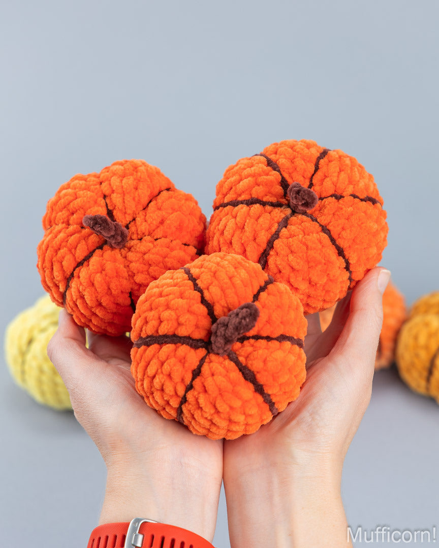 Three orange crocheted amigurumi pumpkins held in two hands against a light gray background, with additional colorful pumpkins blurred in the background and a Mufficorn! logo in the bottom right corner.