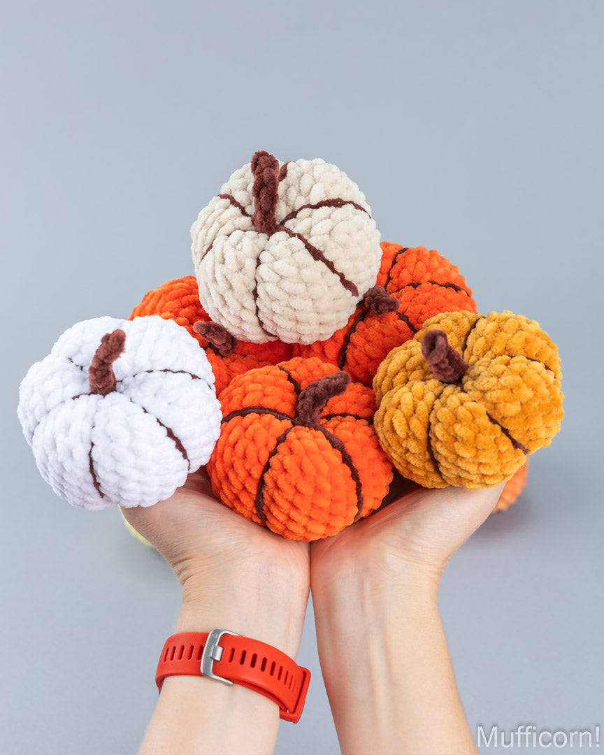 A pair of hands holding crochet plush decorative pumpkins in orange, cream, and mustard yellow, with dark brown stems, against a plain light gray background.