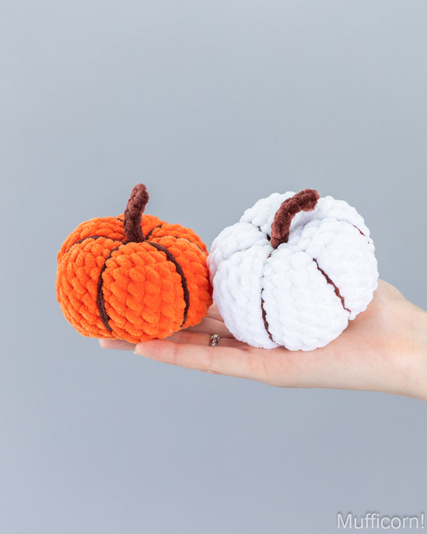 Two amigurumi pumpkins, one orange with dark brown stripes and stem, and one white with dark brown stripes and stem, held in a person's hand against a solid gray background.