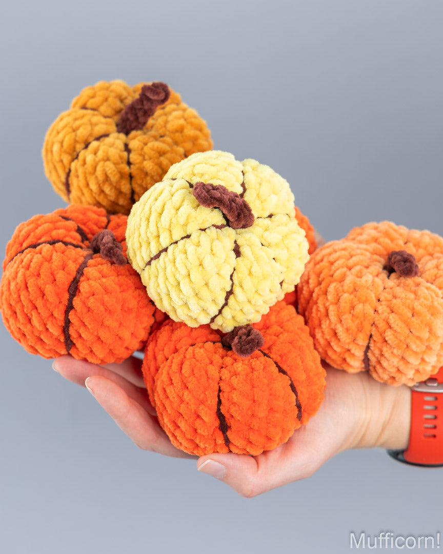 A hand holds a cluster of five small, crochet plush, textured decorative pumpkins in orange, yellow, and mustard tones with dark brown stems, set against a solid gray background.