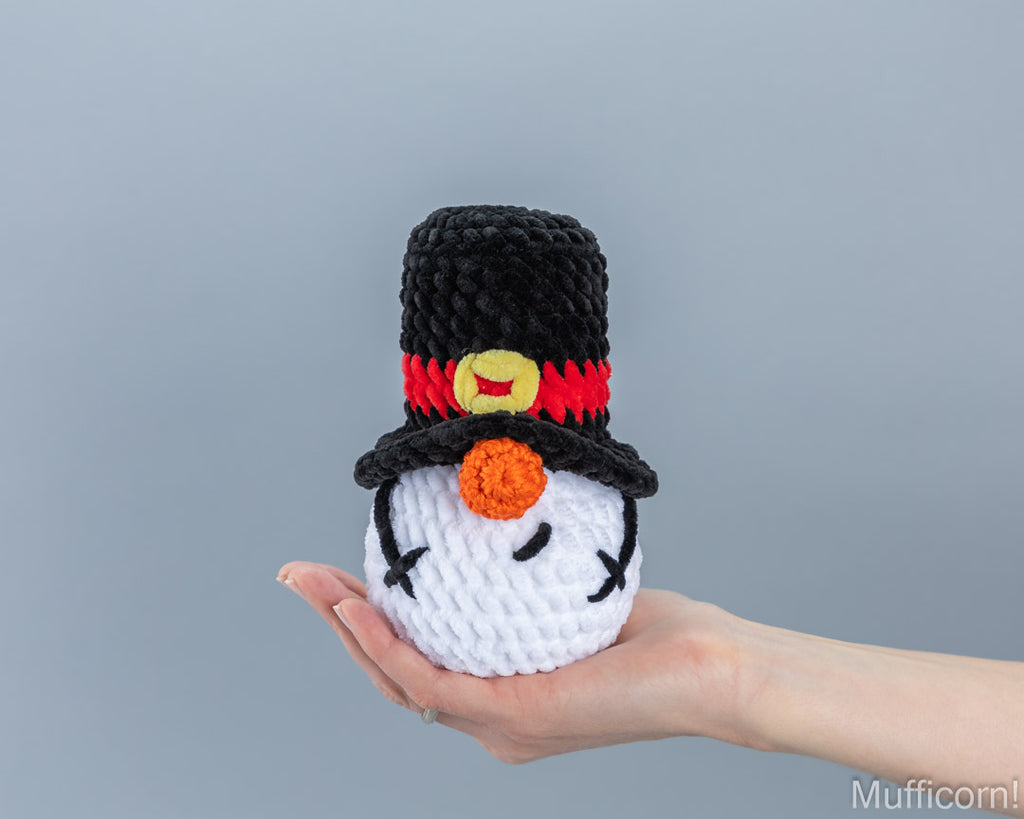 A crocheted snowman figure with a black top hat, red band, yellow button, and orange carrot nose is being held in a person's hand against a plain gray background.