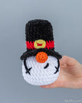 A handmade crocheted snowmangnome in a black top hat with a red band and yellow button, held in a person's hand against a plain gray background.