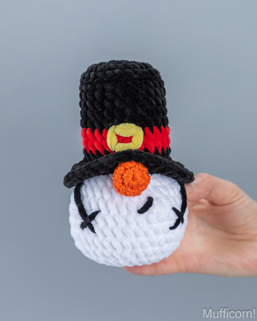 A handmade crocheted snowmangnome in a black top hat with a red band and yellow button, held in a person's hand against a plain gray background.