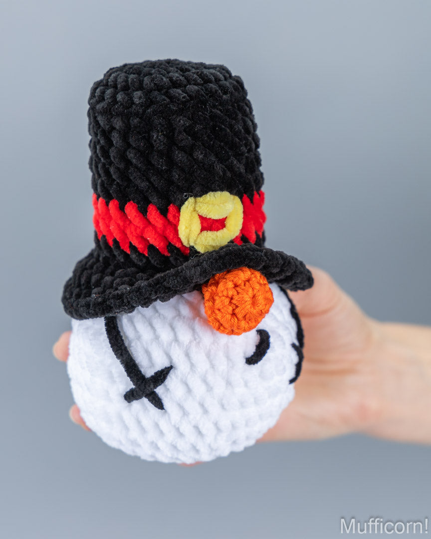 A hand holding a crocheted snowman gnome wearing a black top hat with a red band and yellow emblem, against a plain gray background.