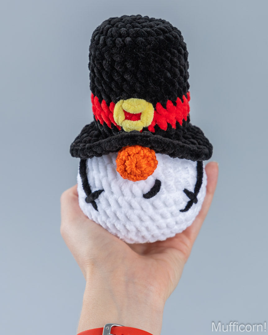 A hand holds a crocheted snowman gnome wearing a black top hat with a red band and yellow button, against a plain light gray background.