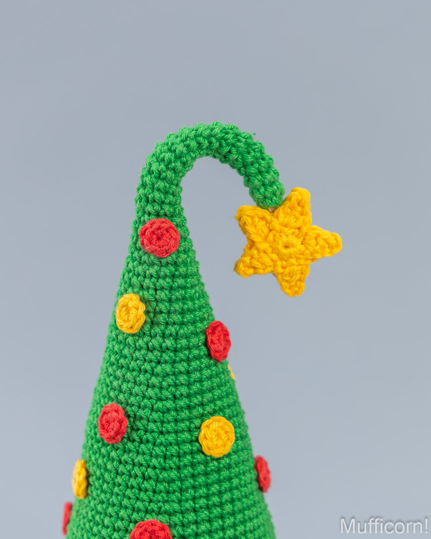 Close-up of a crocheted Christmas tree decoration with a green cone shape, yellow star topper, and red and yellow decorative accents on a light gray background.