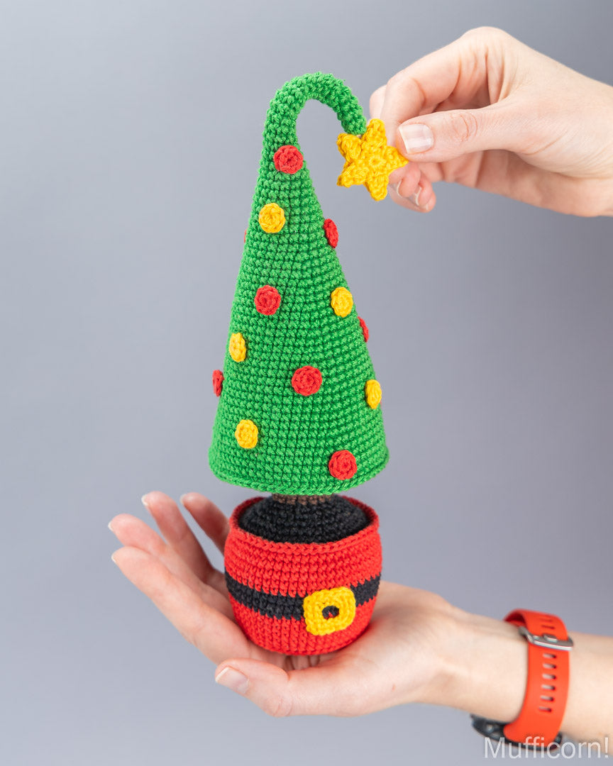 A crocheted Christmas tree hat decoration with a green cone, red and yellow pom-poms, a yellow star topper, and a red-and-black Santa-style base, being held in two hands against a plain gray background.