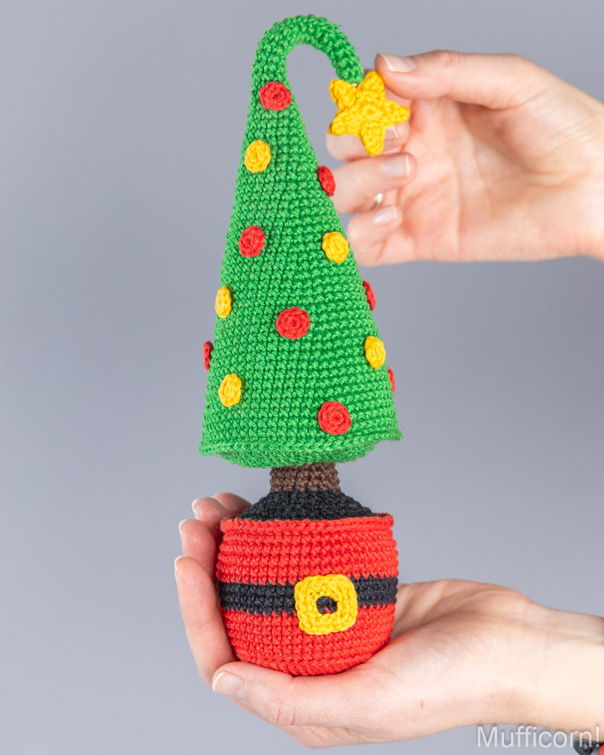 A crocheted Christmas tree ornament with a green cone-shaped tree, red and yellow ornaments, a brown trunk, and a red Santa-style base with black and yellow belt details, being held in two hands against a gray background.