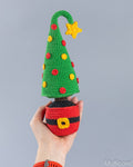 A hand holds a crocheted Christmas tree ornament shaped like a Santa hat, with a green tree, red and yellow ornaments, a yellow star, and a red Santa hat base against a plain gray background.