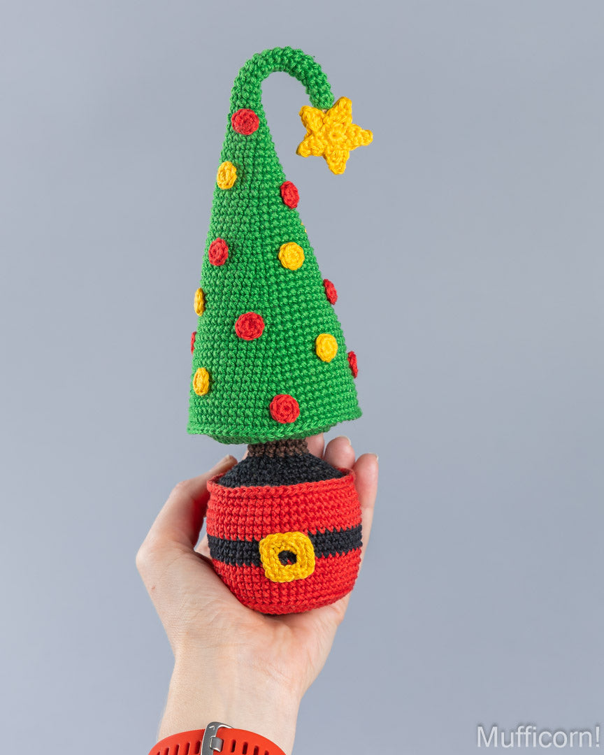 A hand holds a crocheted Christmas tree ornament shaped like a Santa hat, with a green tree, red and yellow ornaments, a yellow star, and a red Santa hat base against a plain gray background.