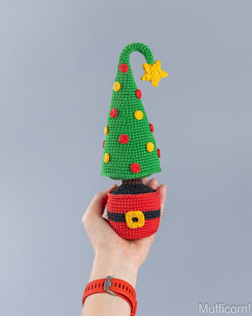 A hand holds a crocheted Christmas tree ornament shaped like a Santa hat, with a green tree, red and yellow ornaments, a yellow star, and a red-and-black hat base, against a plain gray background.