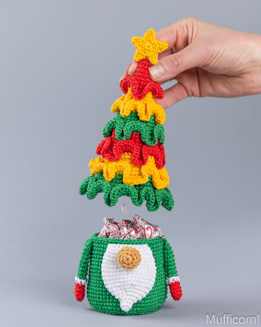 A crocheted Christmas elf gift bag filled with candy, with a hand holding a crocheted Christmas tree topper above it against a plain gray background.