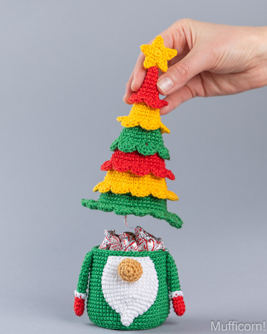 A hand holds a crocheted Christmas tree lid with a yellow star on top, which fits into a green gnome-shaped candy bag filled with wrapped candies, against a solid gray background.