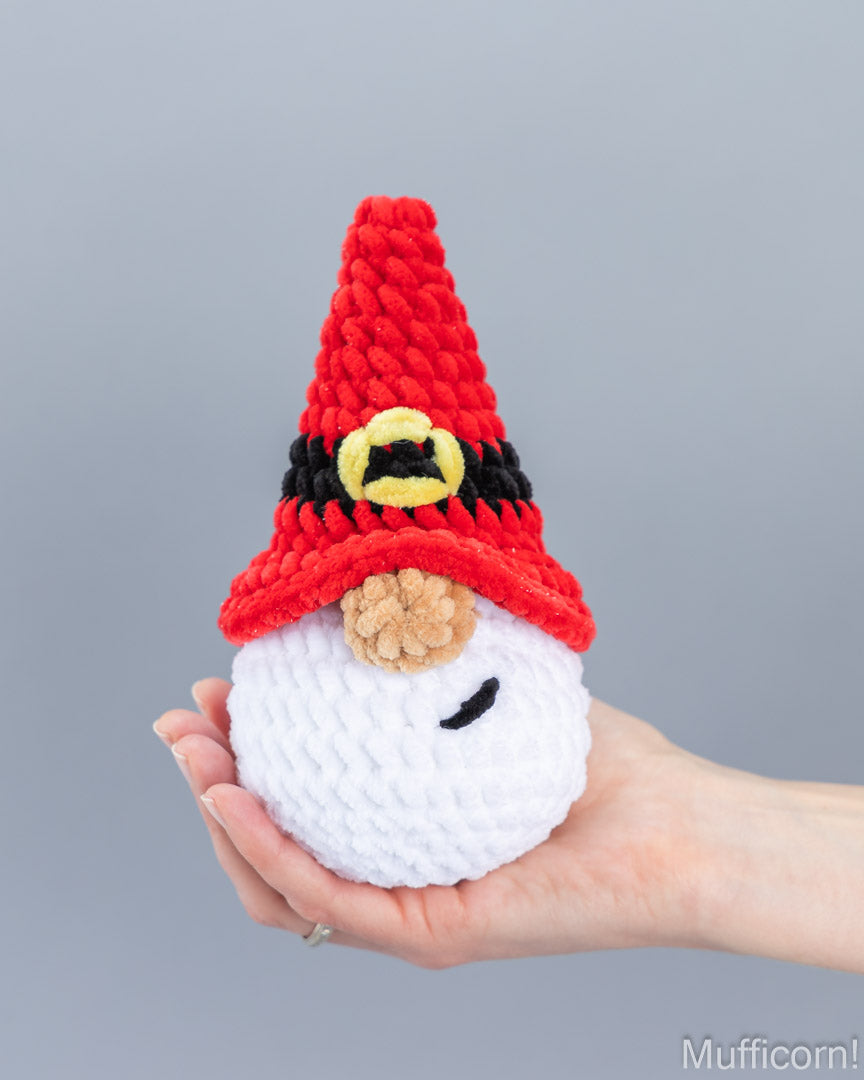 A hand holding a small crocheted plush santa gnome with a red pointed hat, black and yellow band, tan nose, and white body against a plain gray background.
