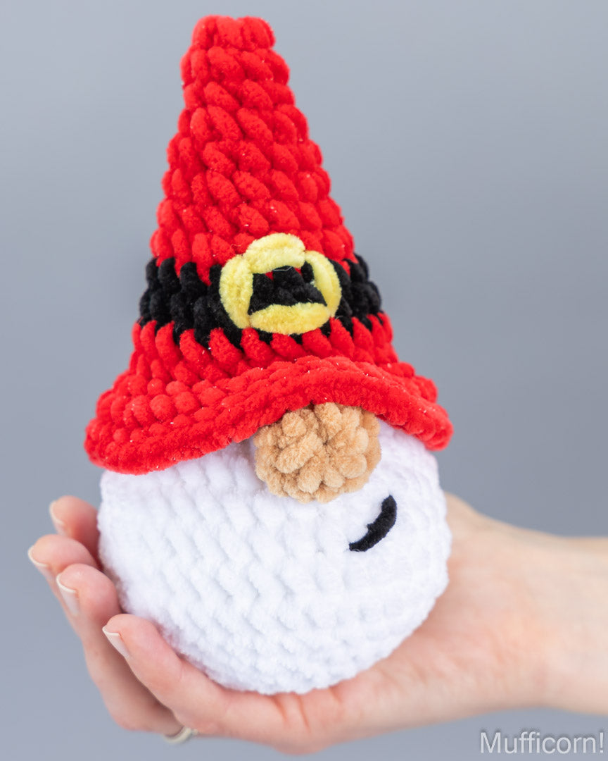 A hand holding a small crocheted santa gnome toy with a red pointed hat, black and yellow band, tan nose, and white body against a plain gray background.
