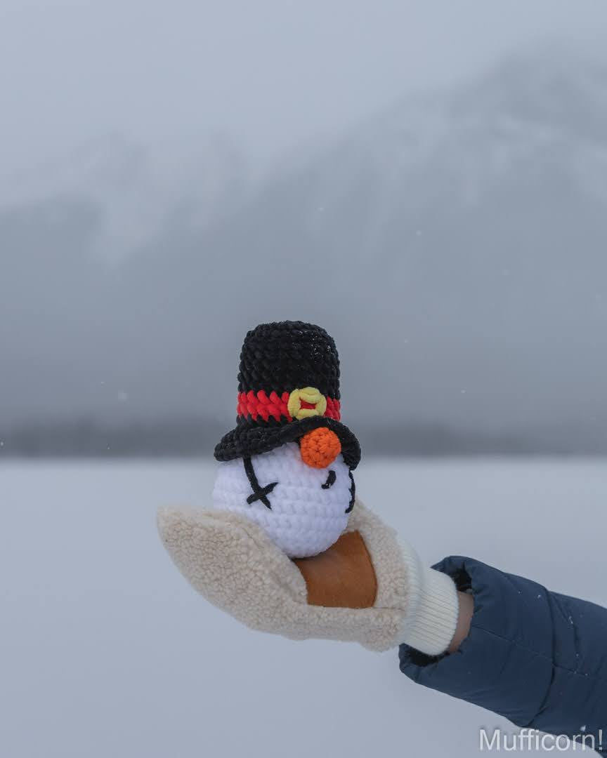 A crocheted snowman figure held in a hand wearing a blue puffer jacket and beige winter glove, with a snowy outdoor landscape and blurred mountains in the background.