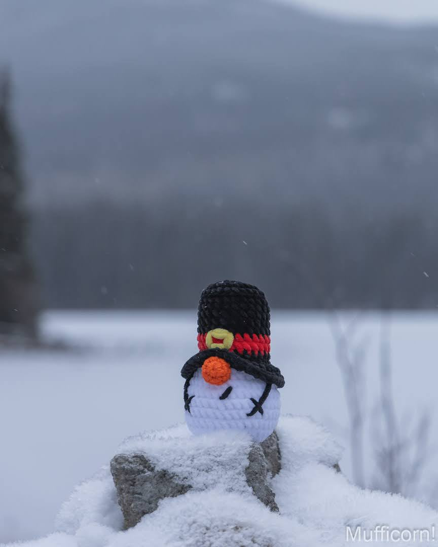 A small crocheted snowman gnome wearing a black top hat with a red band and yellow buckle, standing outdoors in snow with a blurred winter landscape in the background.