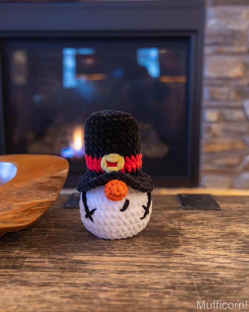 A small crocheted snowman gnome ornament sits on a wooden table in front of a fireplace. The snowman has a white body, black hat, red and yellow band, and orange carrot nose.