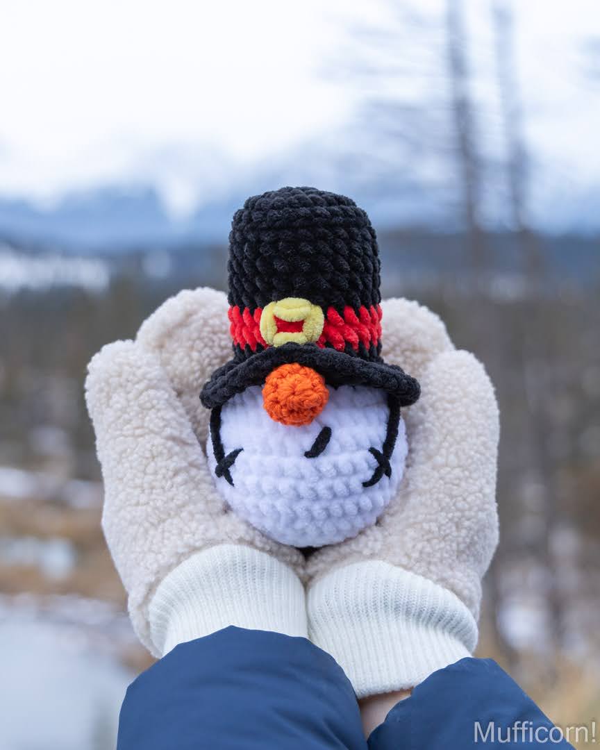 A crocheted snowman plush gnome held in cream-colored fleece gloves, with a blurred outdoor winter landscape in the background. The accessory features a black top hat with red and yellow accents, a white face, and an orange carrot nose.
