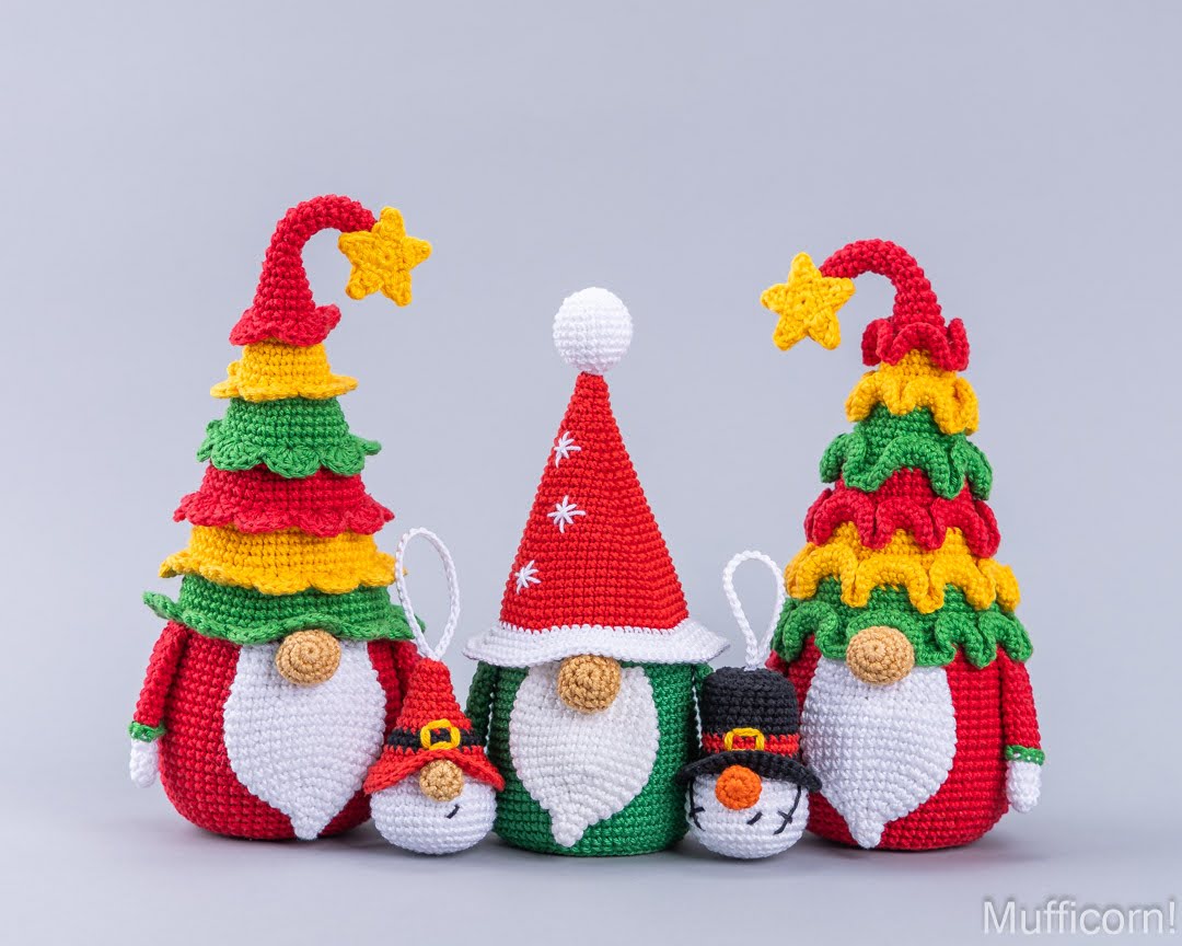 Three crocheted Christmas gnome figures with colorful tree-shaped hats, shown on a light gray studio background. The center gnome wears a red Santa hat with white trim and white beard, flanked by two gnomes with red, green, and yellow tree hats and white beards.