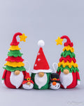 Four crocheted Christmas-themed gnomes in red, green, yellow, and white, arranged on a light gray background. The set includes two larger gnomes with decorated tree hats, one small gnome with a Santa hat, and one snowman.