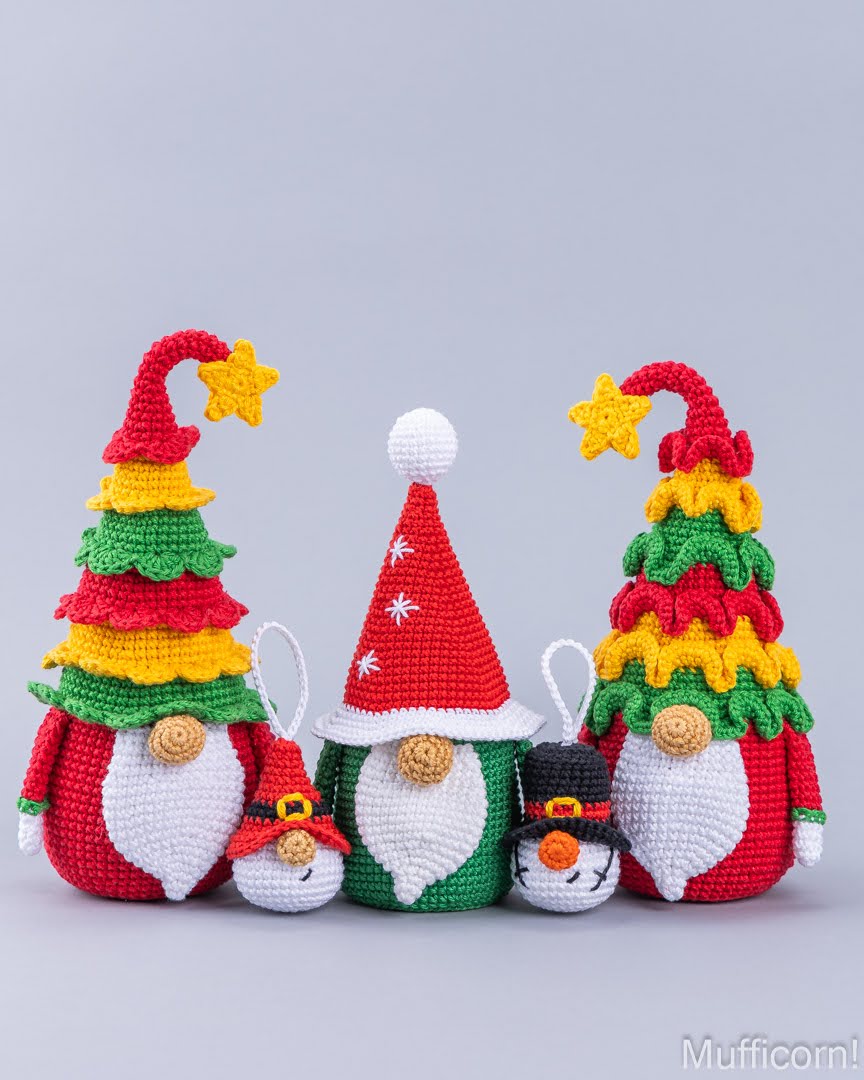 Four crocheted Christmas-themed gnomes in red, green, yellow, and white, arranged on a light gray background. The set includes two larger gnomes with decorated tree hats, one small gnome with a Santa hat, and one snowman.
