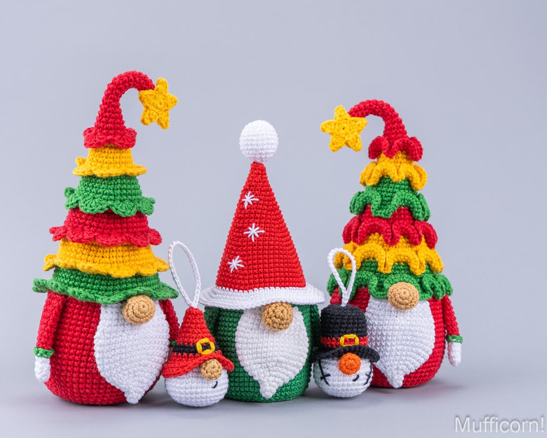 Four crocheted Christmas gnomes with colorful tree-shaped hats, shown on a light gray studio background. The gnomes have red, green, white, yellow, and black details, with two wearing Santa hats and two wearing tree hats decorated with stars.