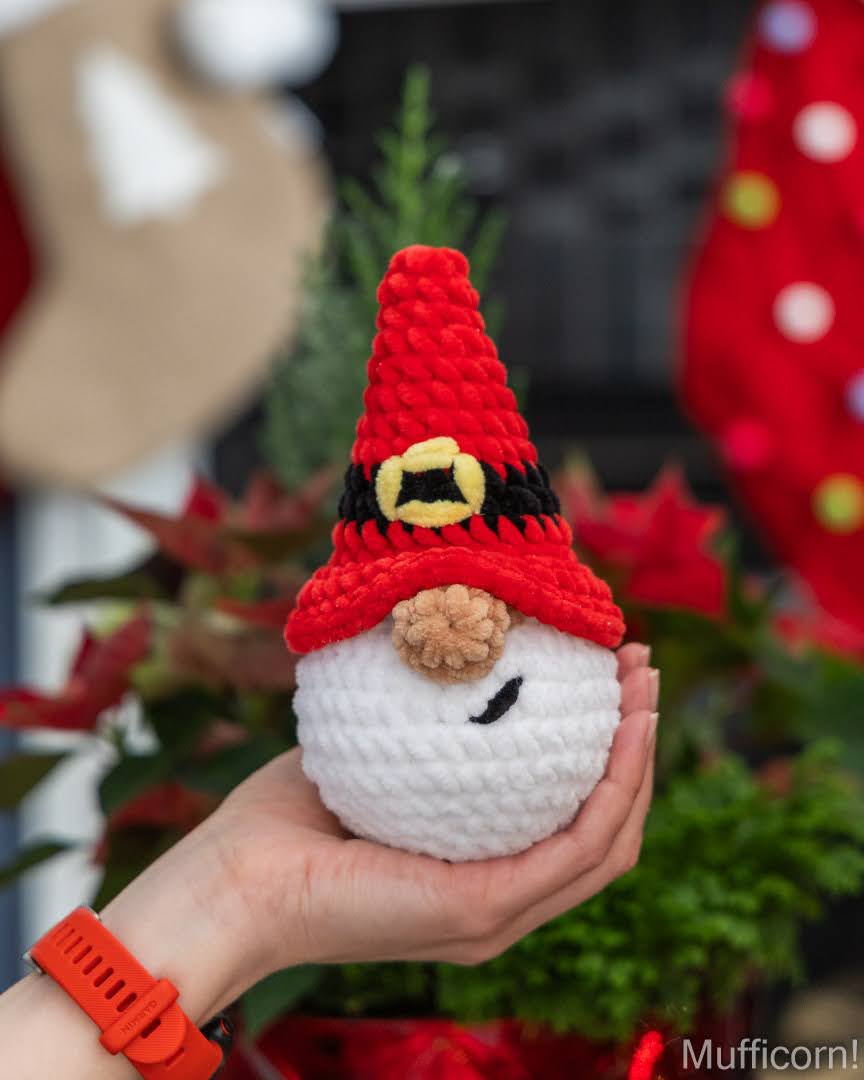 A hand holds a small crocheted plush santa gnome ornament wearing a red pointed hat with a black band and yellow buckle detail, with blurred holiday decorations in the background.