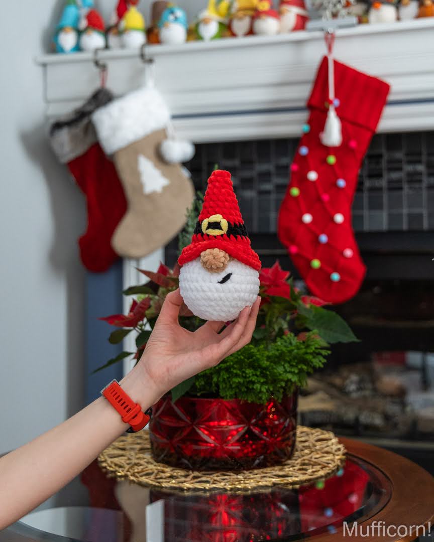 A hand holds a crocheted white Santa gnome  ornament wearing a red pointed hat with a gold bell detail, set against a cozy holiday background with stockings, a fireplace, and a potted plant.