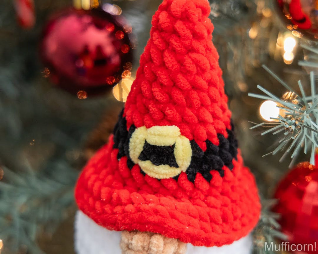 A close-up of a red yarn Christmas Santa gnome ornament wearing a black and yellow hat, set against a blurred Christmas tree background with red ornaments and lights.