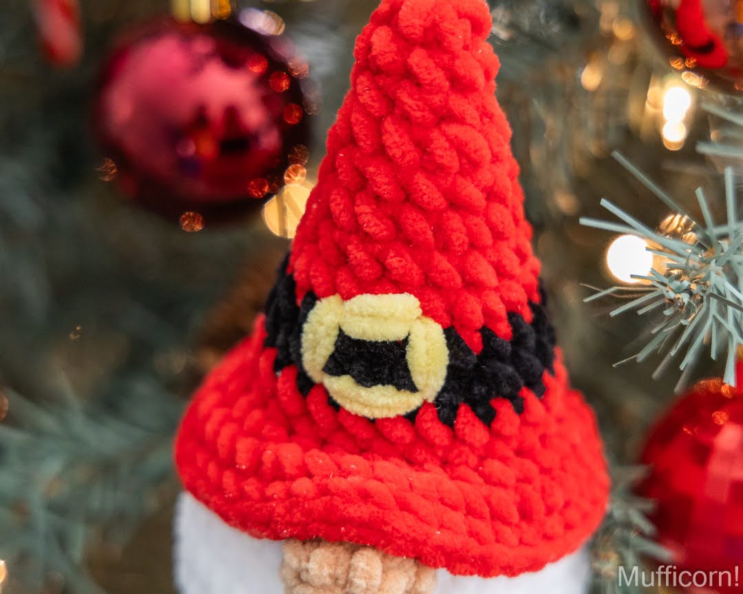 A close-up of a red yarn Christmas Santa gnome ornament wearing a black and yellow hat, set against a blurred Christmas tree background with red ornaments and lights.