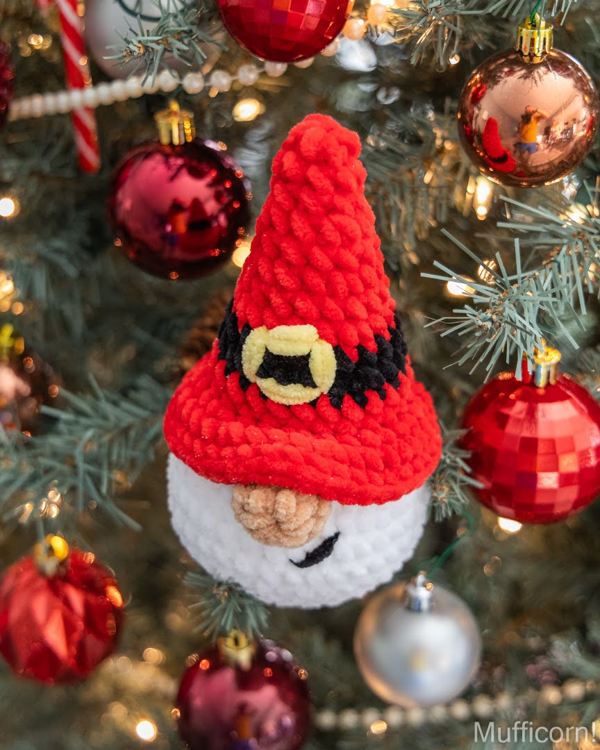 A crocheted Christmas Santa gnome ornament hanging on a decorated Christmas tree. The ornament has a red pointed hat with a black band and yellow buckle detail, a white face, and a beige stitched nose.