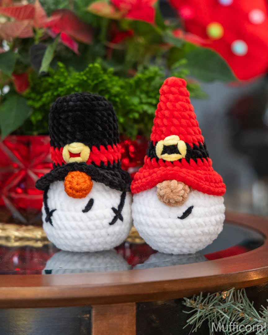 Two crocheted snowman ornaments sitting on a reflective wooden table, decorated with black and red hats, colorful scarves, and white bodies, with festive greenery and red flowers blurred in the background.