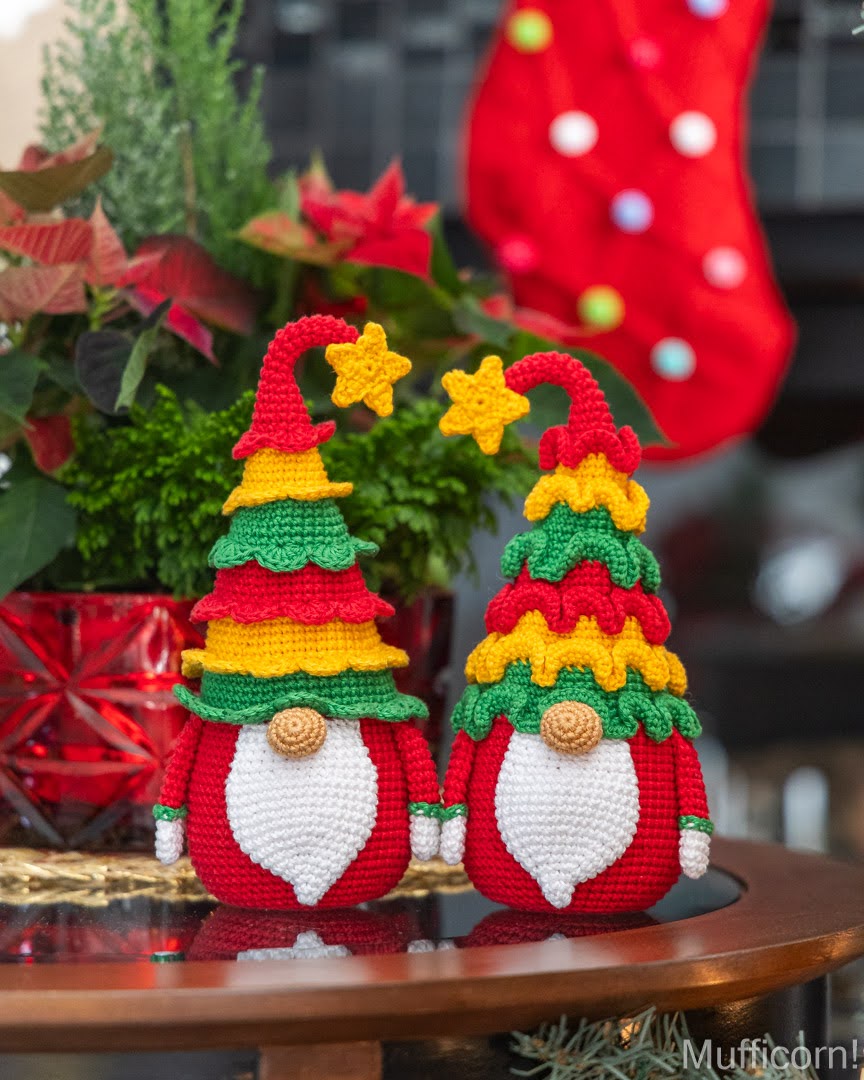 Two crocheted Christmas gnome figures styled as tree-shaped hats, sitting on a wooden table with holiday plants and a red stocking in the background.