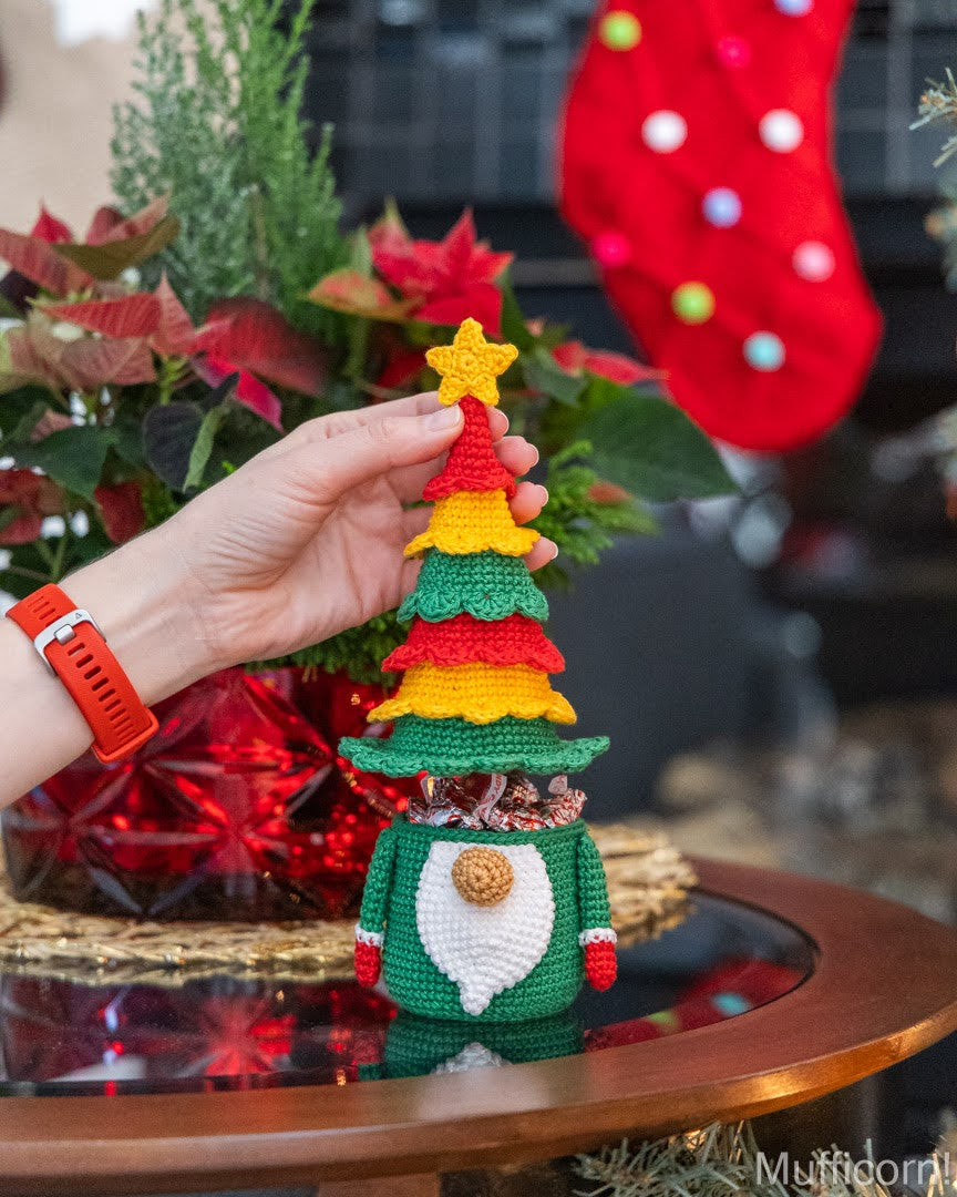 A crocheted Christmas gnome figurine with a tiered green, red, and yellow tree hat, being held by a hand. The gnome has a white beard, green body, and red hands, set against a festive holiday background with poinsettias and a red stocking.