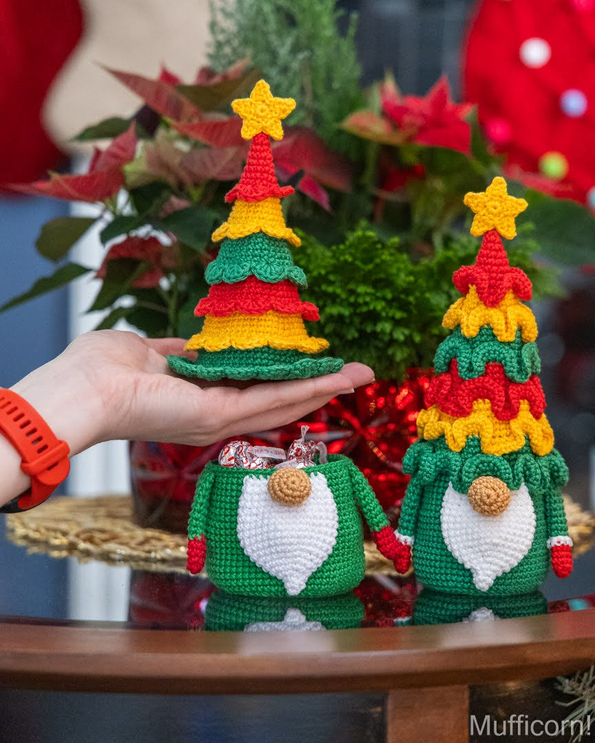 Two crocheted Christmas tree gnomes displayed on a wooden table, with one held in a person's hand. The background features festive holiday decorations including poinsettias and a red stocking.