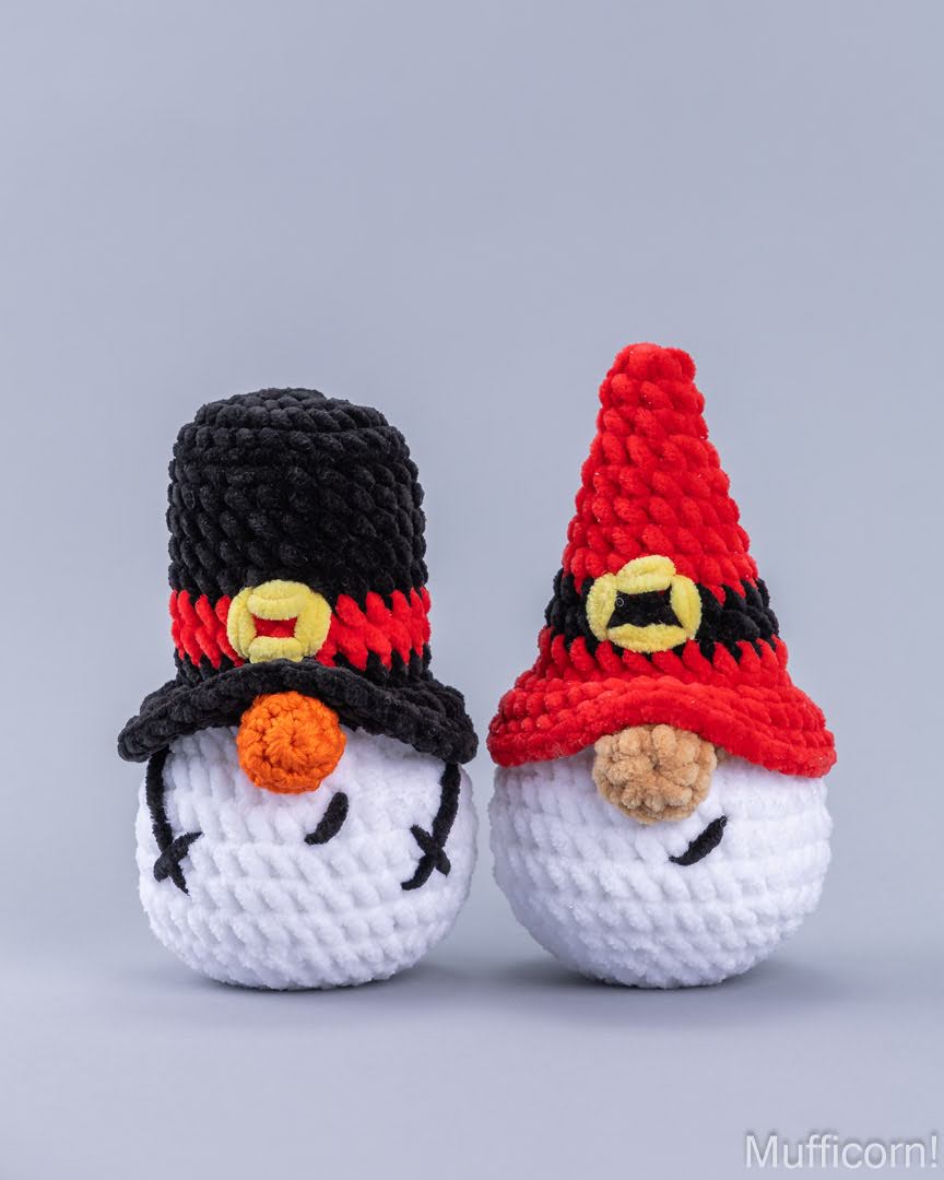Two crocheted snowman toys side by side on a light gray background. One snowman wears a black hat with a red band and yellow buckle, and the other wears a red hat with a black band and yellow buckle.