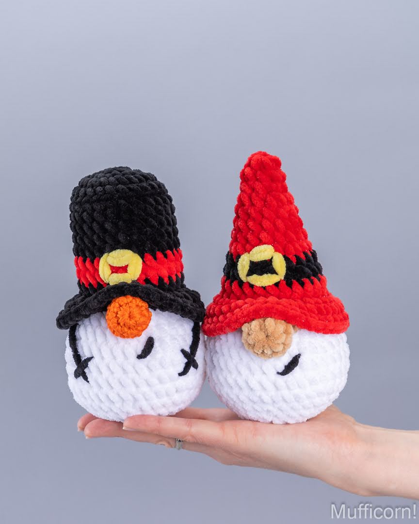 Two crocheted gnome figures are held in a person's hand against a plain gray background. One gnome wears a black hat with a red band and yellow medallion, and the other wears a red hat with a black band and yellow medallion.